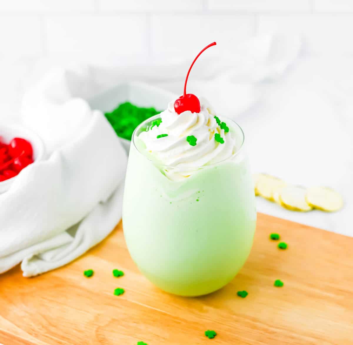 Shamrock shake on wood board with green sprinkles, cherries and white towel surround.
