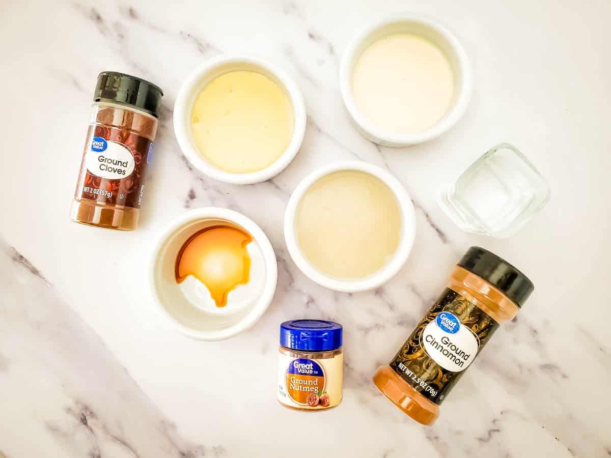 4 white bowls with drink ingredients alongside cinnamon, nutmeg and cloves spice bottles