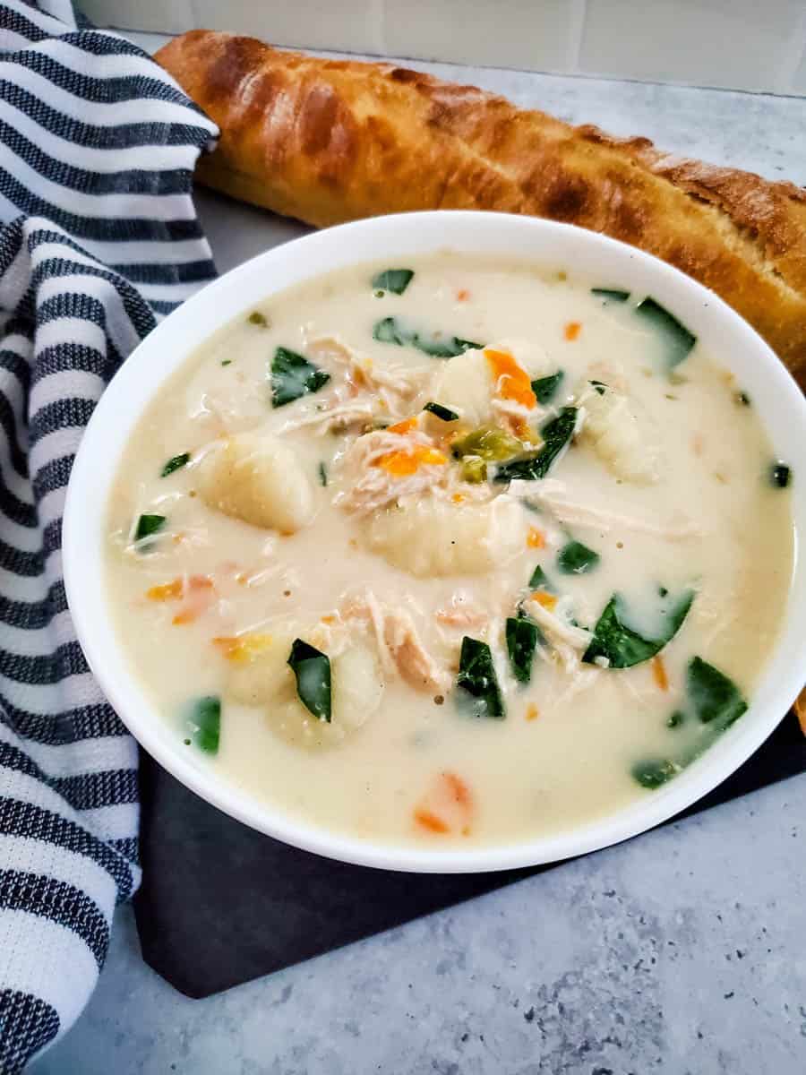 Bowl of creamy chicken gnocchi soup next to blue and white strip towel with bread in background.