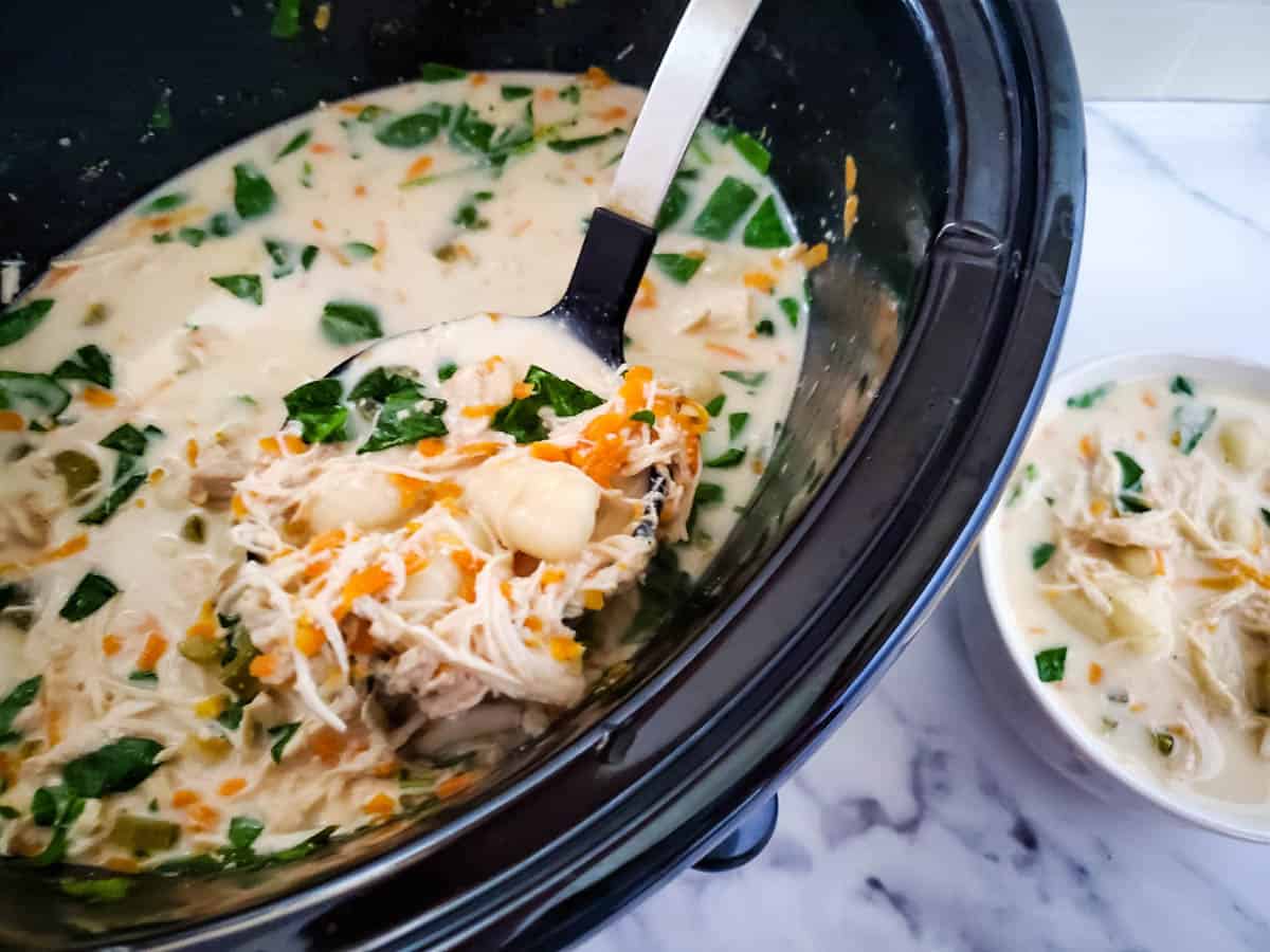 Slow cooker with finished soup and ladle lifting soup out of pot. Bowl of soup next to slow cooker.