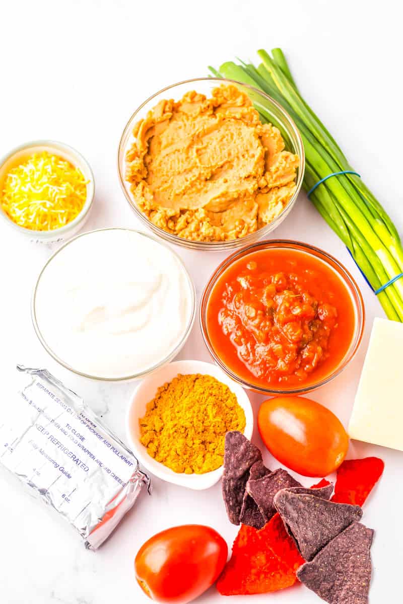 All ingredients in bowls for taco dip with tomatoes, green onions and blue and red chips on the side