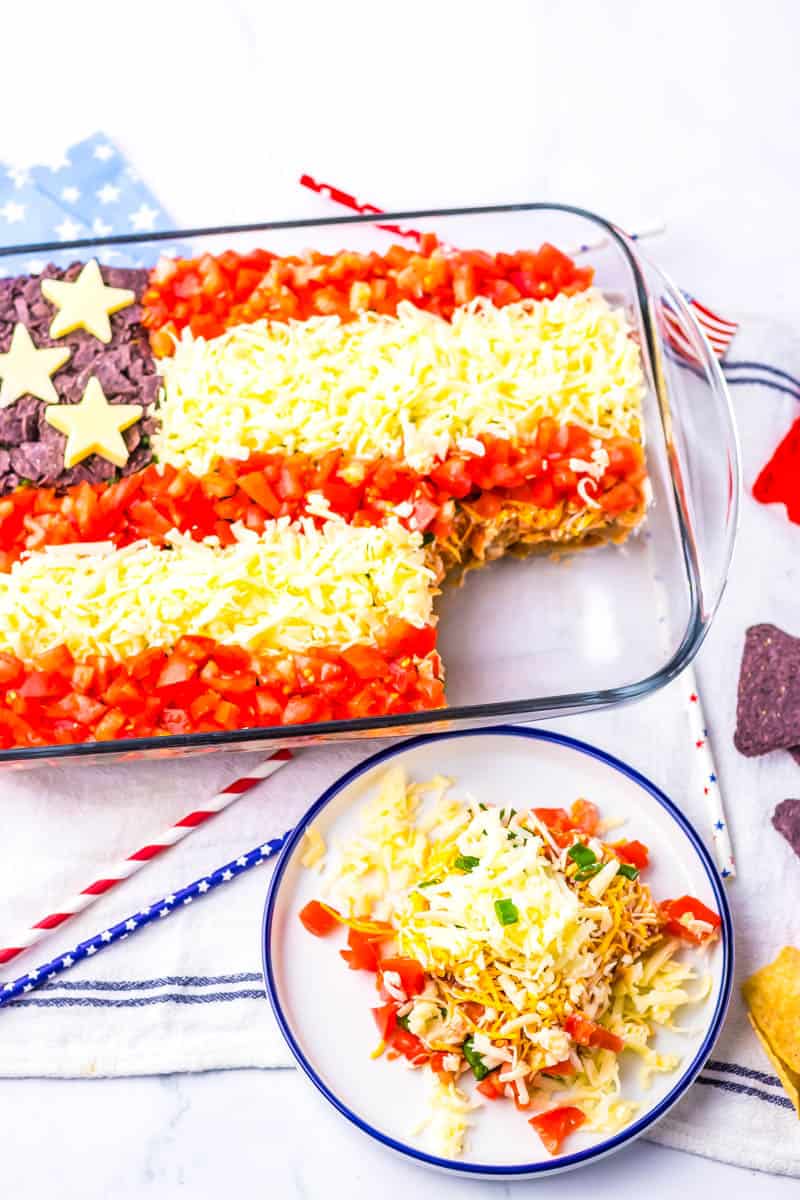Taco dip plated with corner of main dish missing surrounding by patriotic straws and blue and red chips.