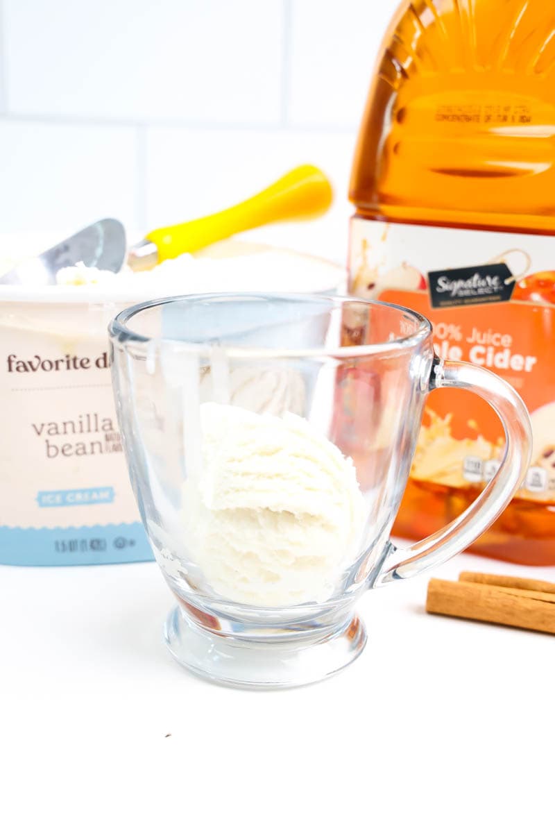 Clear glass with a scoop of vanilla ice cream on white backdrop with ice cream and apple cider in the background
