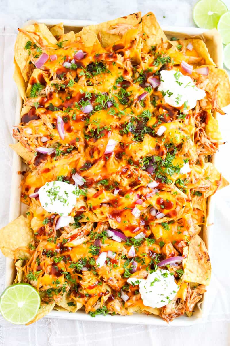 Sheet pan of bbq chicken nachos with sour cream, cheese,, bbq sauce, red onions, parsley and chips.