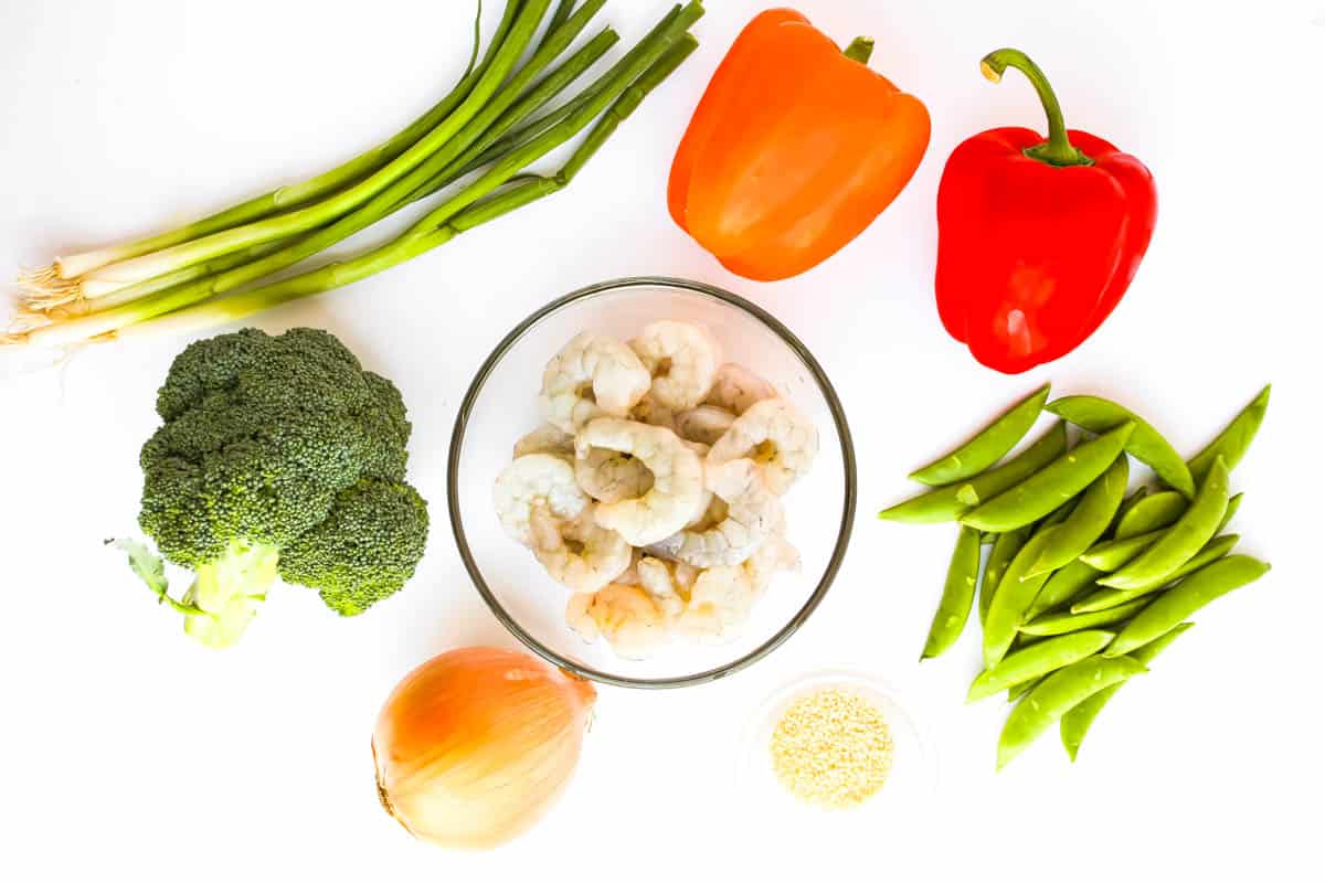 Green onions, red and orange bell pepper, snap peas, broccoli, onion, sesame seeds and shrimp are pictured on white background.