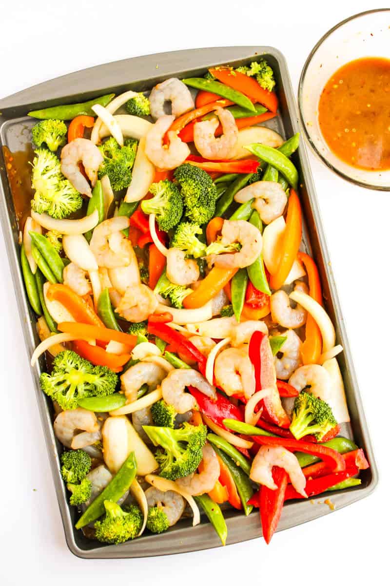 Sheet pan with sliced peppers, onion, broccoli and shrimp with marinade in bowl in the corner on white background.