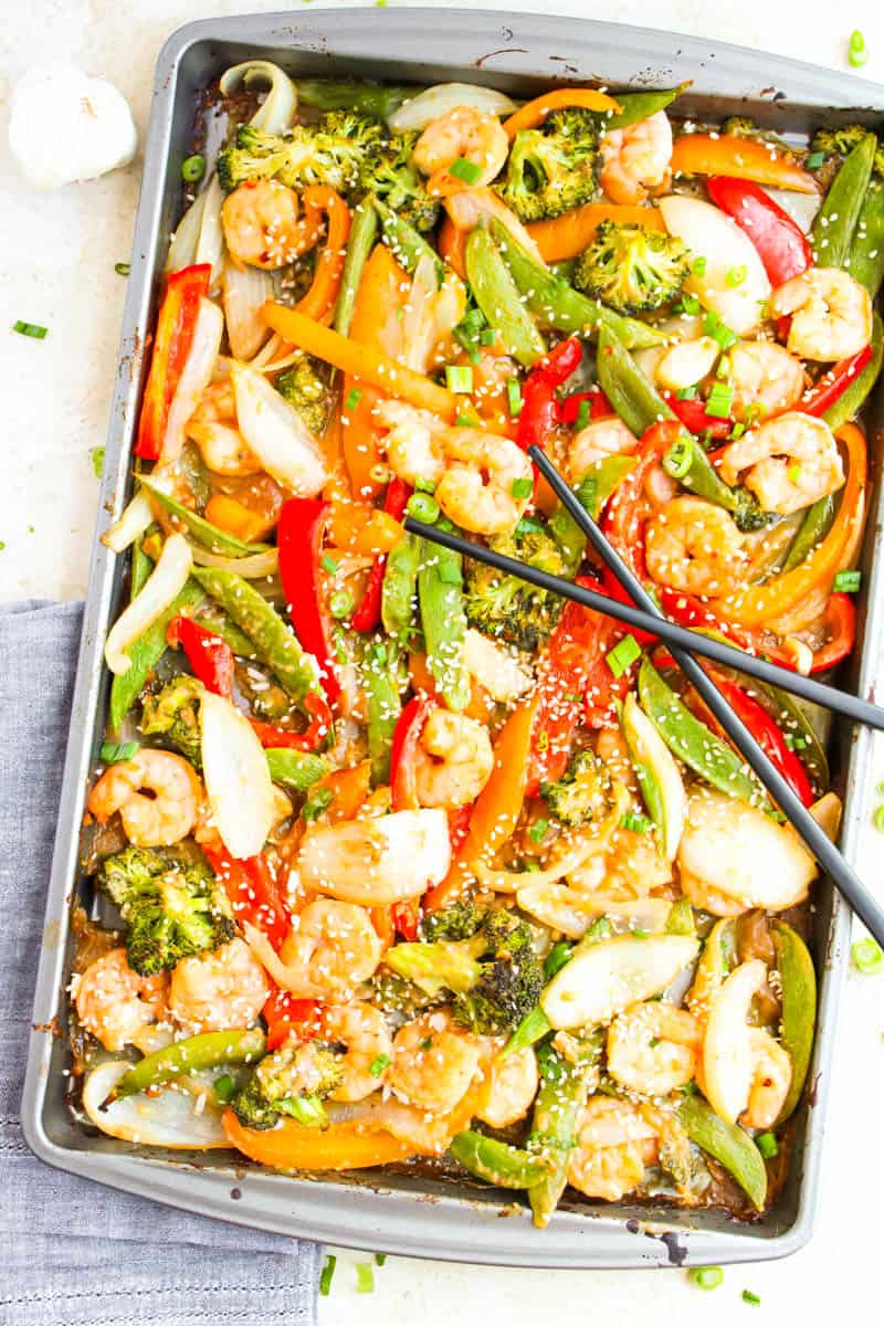 Shrimp and vegetables on one sheet pan with black chopsticks across it. Blue napkin underneath pan on white background.