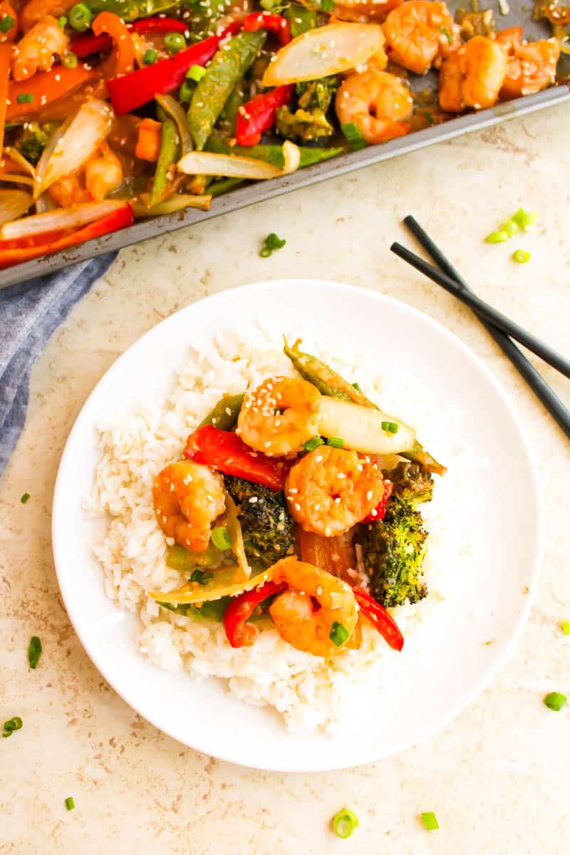Plate with rice and sheet pan ingredients-shrimp, peppers, onion and broccoli on top. Black chopsticks next to plate with sheet pan in the top left corner on white background.
