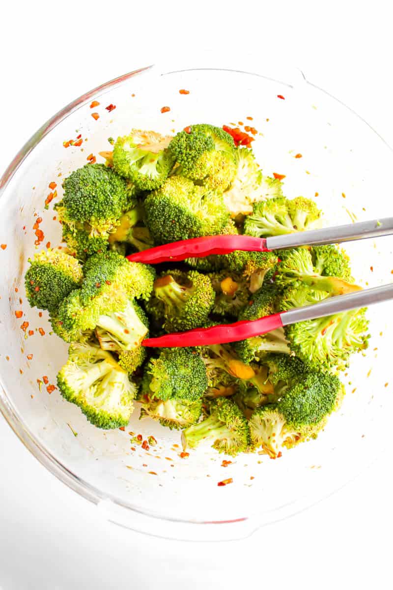 Clean bowl with broccoli in the marinade being tossed with a pair of tongs.