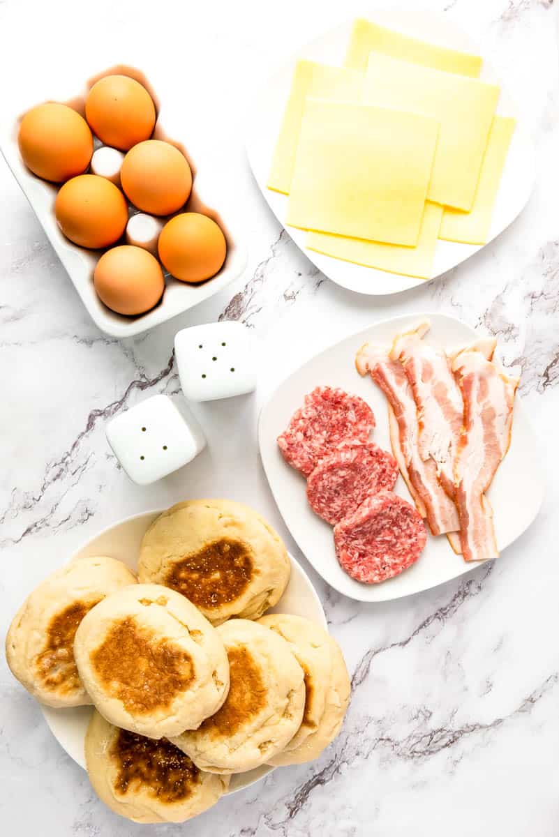 Six eggs, cheese slices on white plate, bacon and sausage patties on white plate, English muffins and salt and pepper shakers on white background.