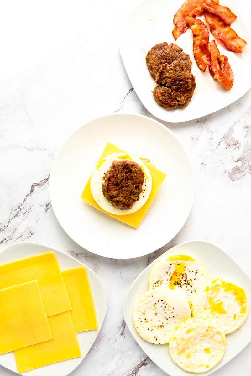 White plate in center with open face sandwich with sausage patty on top. Right corner sausage and bacon. Bottom right egg cups and bottom left corner cheese slices.
