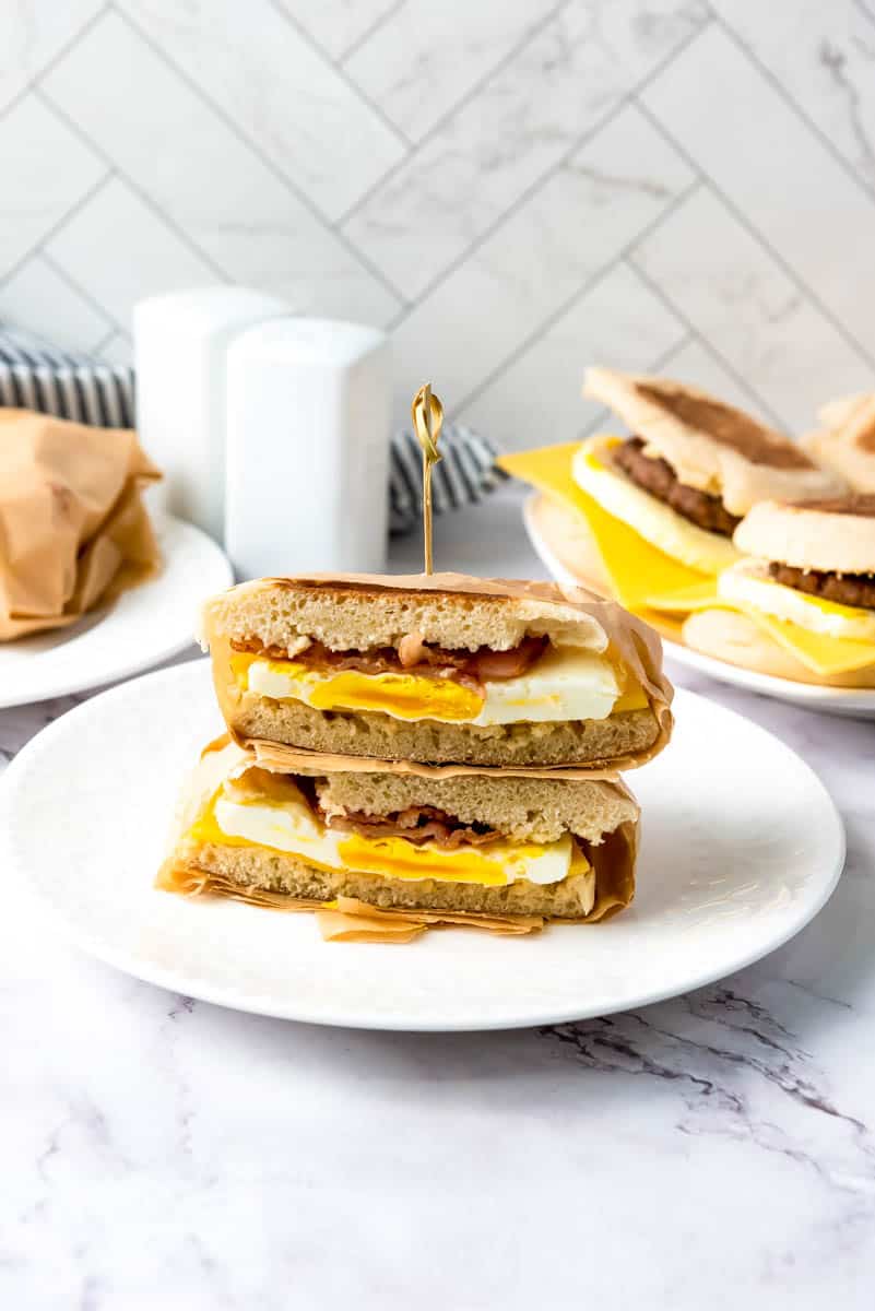 Stacked sandwich on white plate in center with salt and pepper shaker and plate of sandwiches behind on gray marble background.