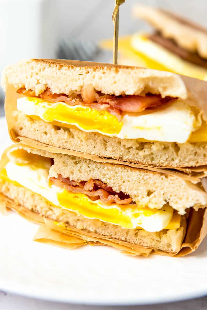 Two sandwiches stacked on top of one another. Bread, egg, bacon and top layer of bread wrapped in parchment paper with toothpick sticking in top.