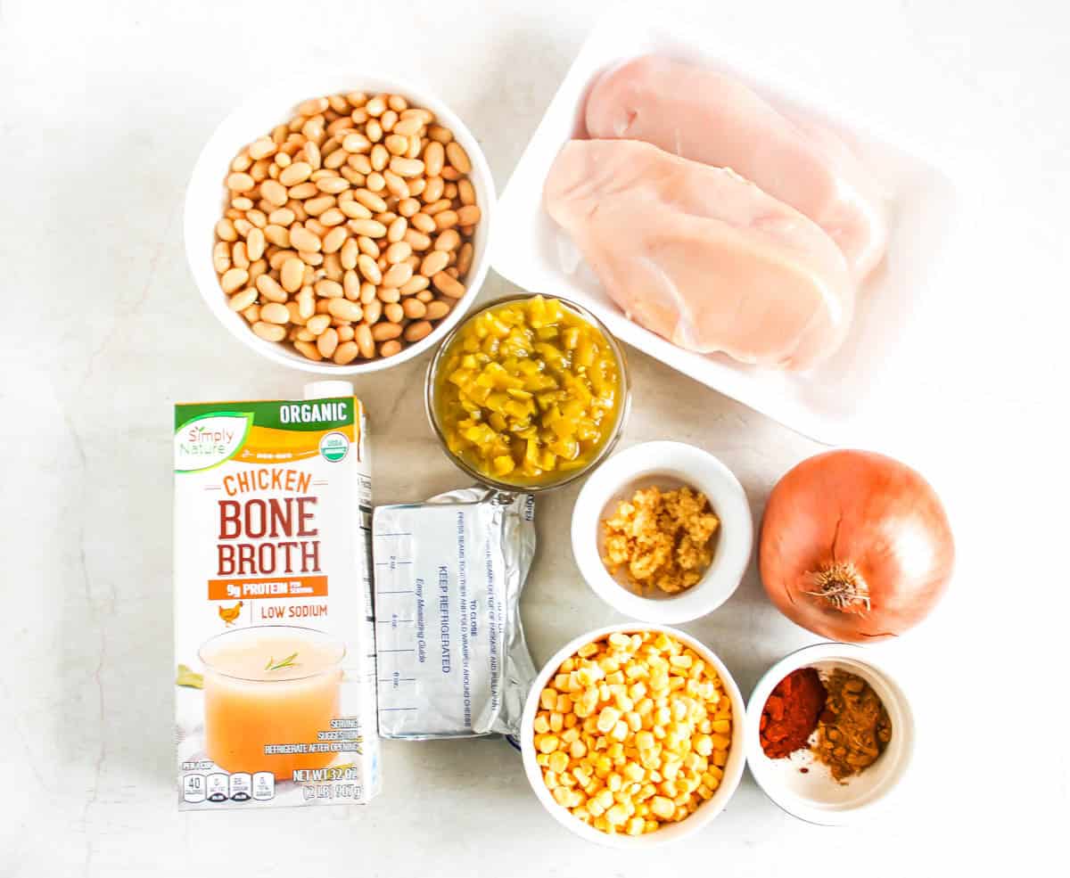 Ingredients for white chicken chili- broth, cream cheese, bowl of beans, cup of green chiles, corn, minced garlic, and spices, along with chicken breast on plate and whole white onion
