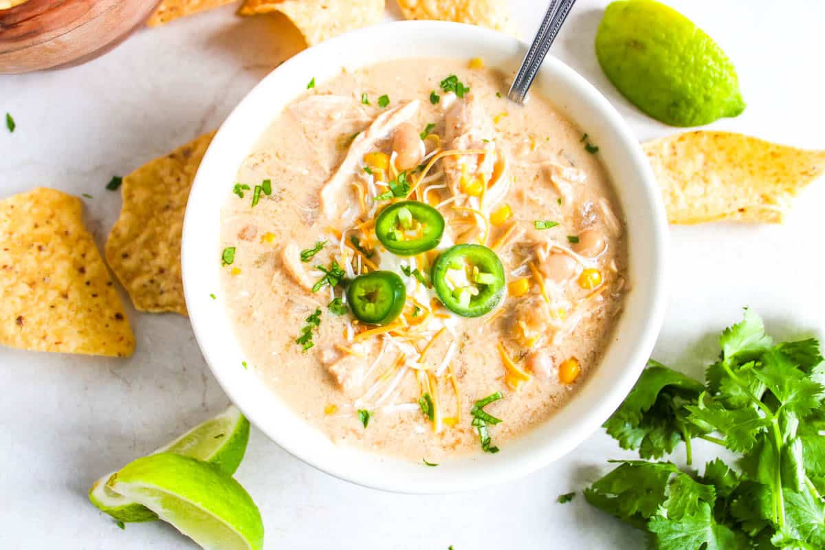 White bowl of white chicken chili surrounded by chips, cilantro and lime wedges.