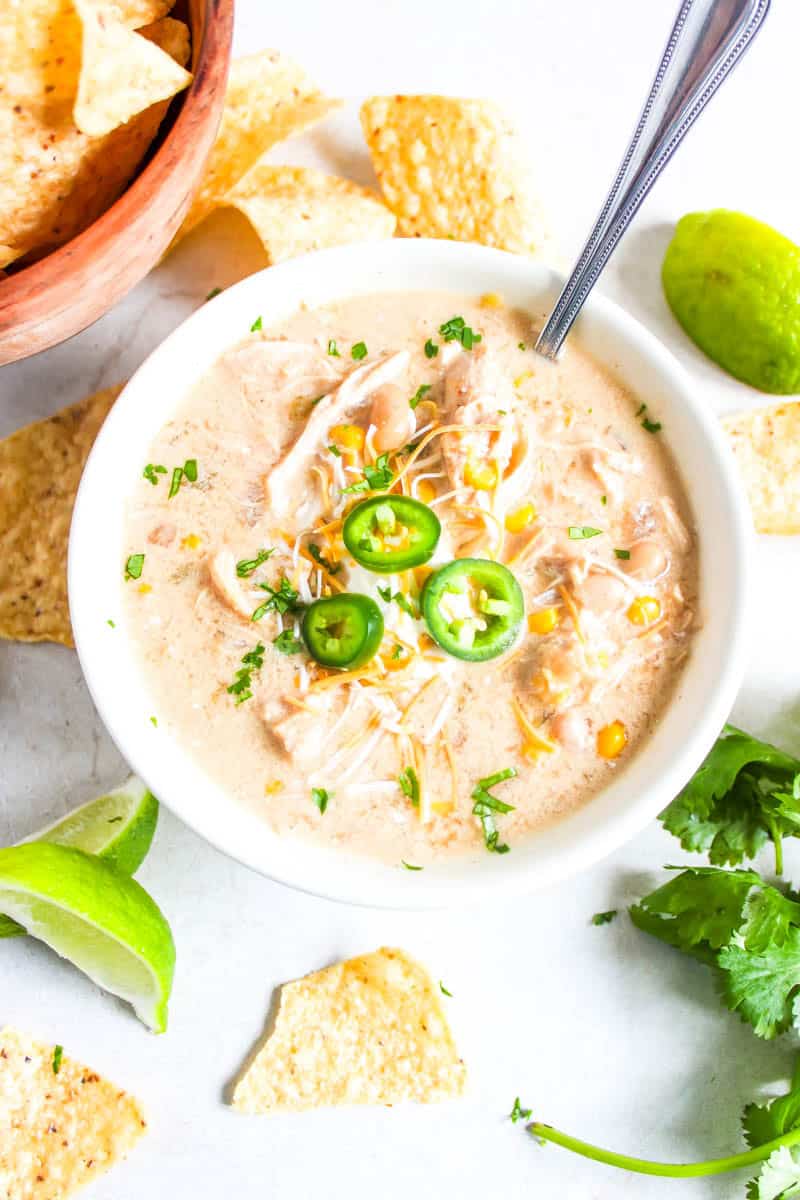 White bowl with white chicken chili surrounded by chip, lime wedges, a lime and cilantro.