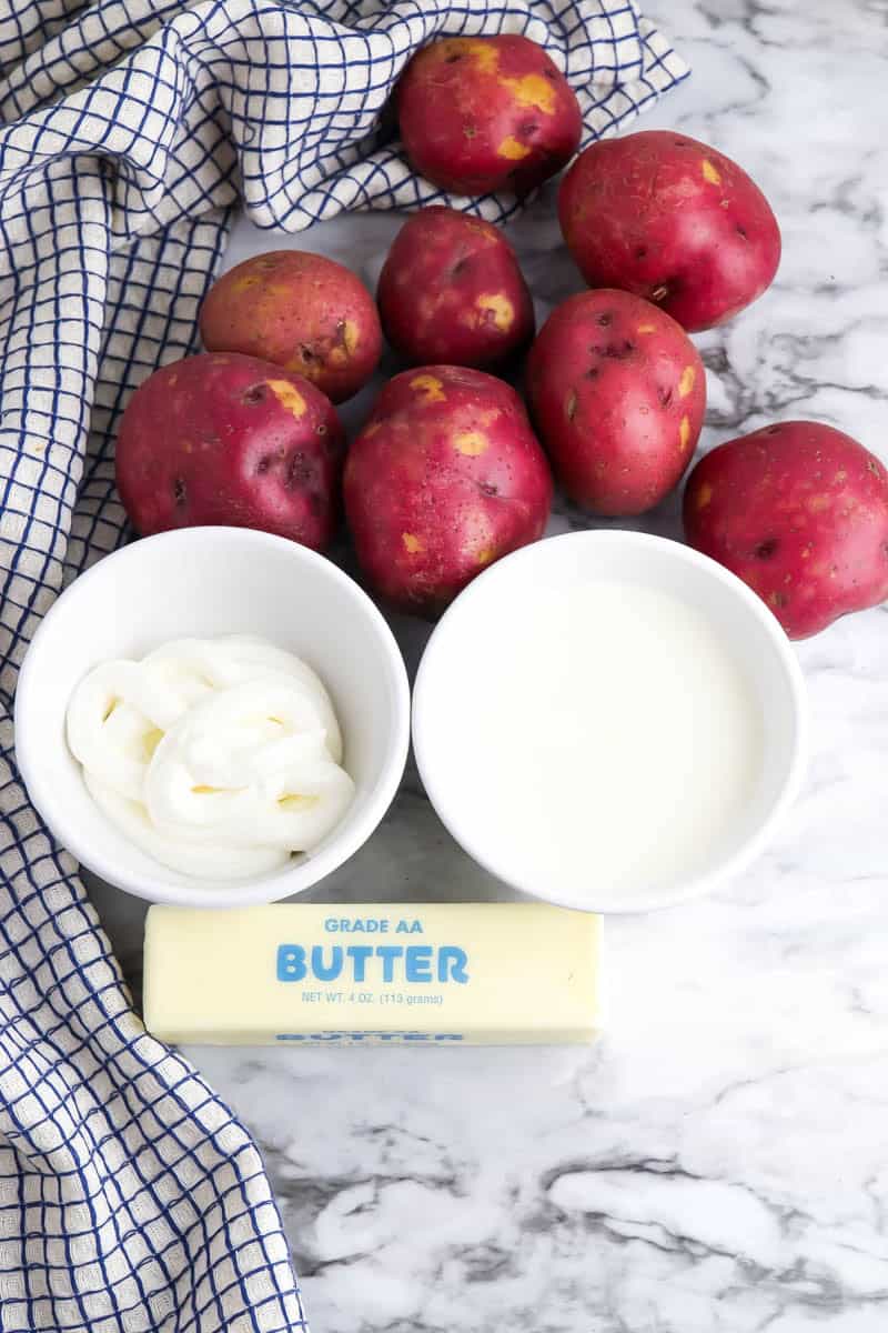 Red skinned potatoes, white bowls with sour cream and heavy cream and a stick of butter on marble background.