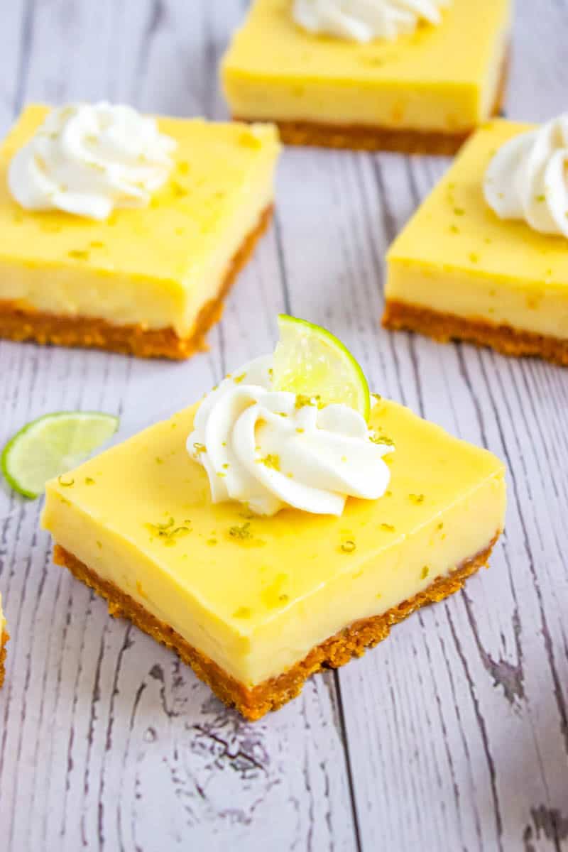 Close up of key lime pie bar on wooden background topped with whipped cream, lime slice and lime zest.