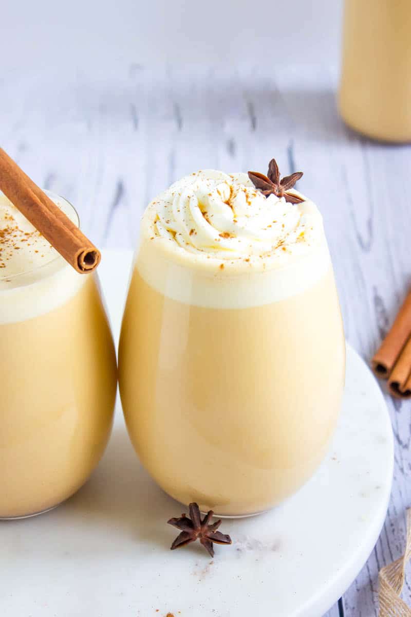 Two pumpkin spice eggnog glasses with cinnamon sticks on a white plate.