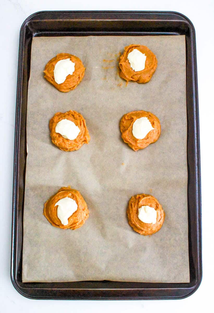 The cookie dough on the parchment paper of the baking sheet with a dollop of cream cheese on top.