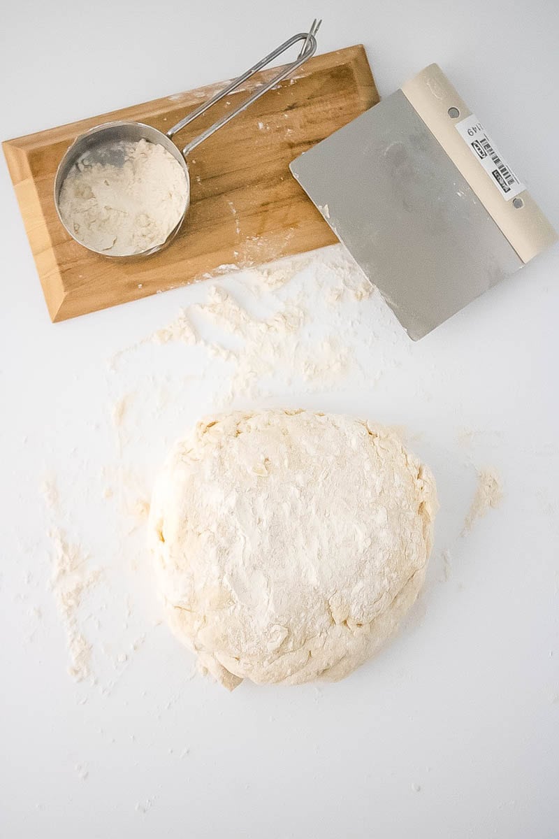A ball of floured dough on white surface with a scraper and metal cookie cutter.