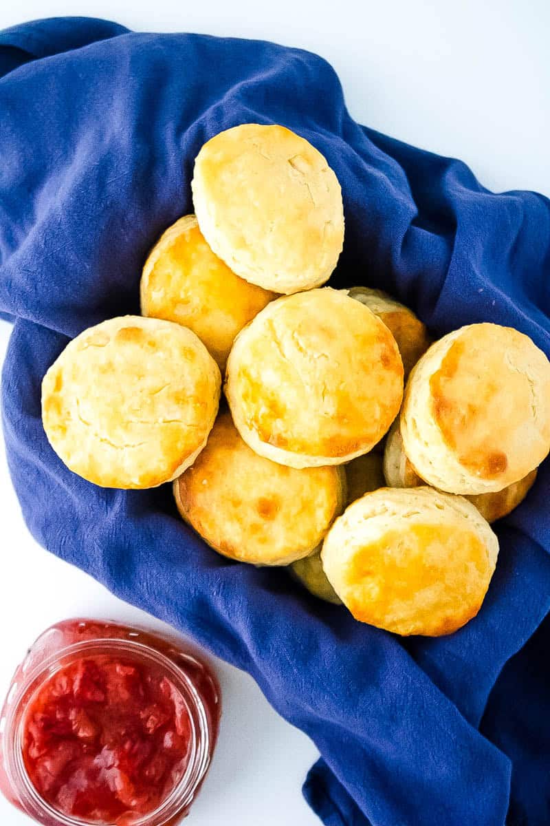 A basket of buttermilk biscuits on a blue towel with a jar of strawberry jam next to it.