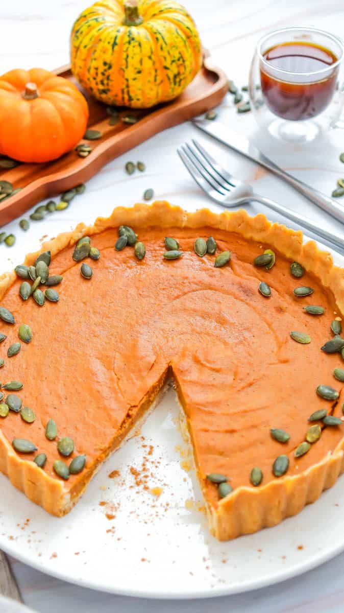 Gluten free pumpkin pie with slice missing on a white background with pumpkin seeds scattered around it.