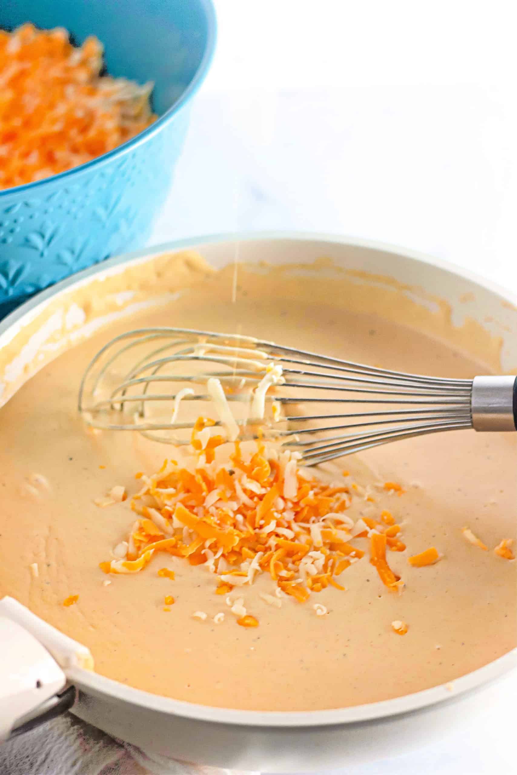Shredded cheese being added and whisked into the roux in a white pan.