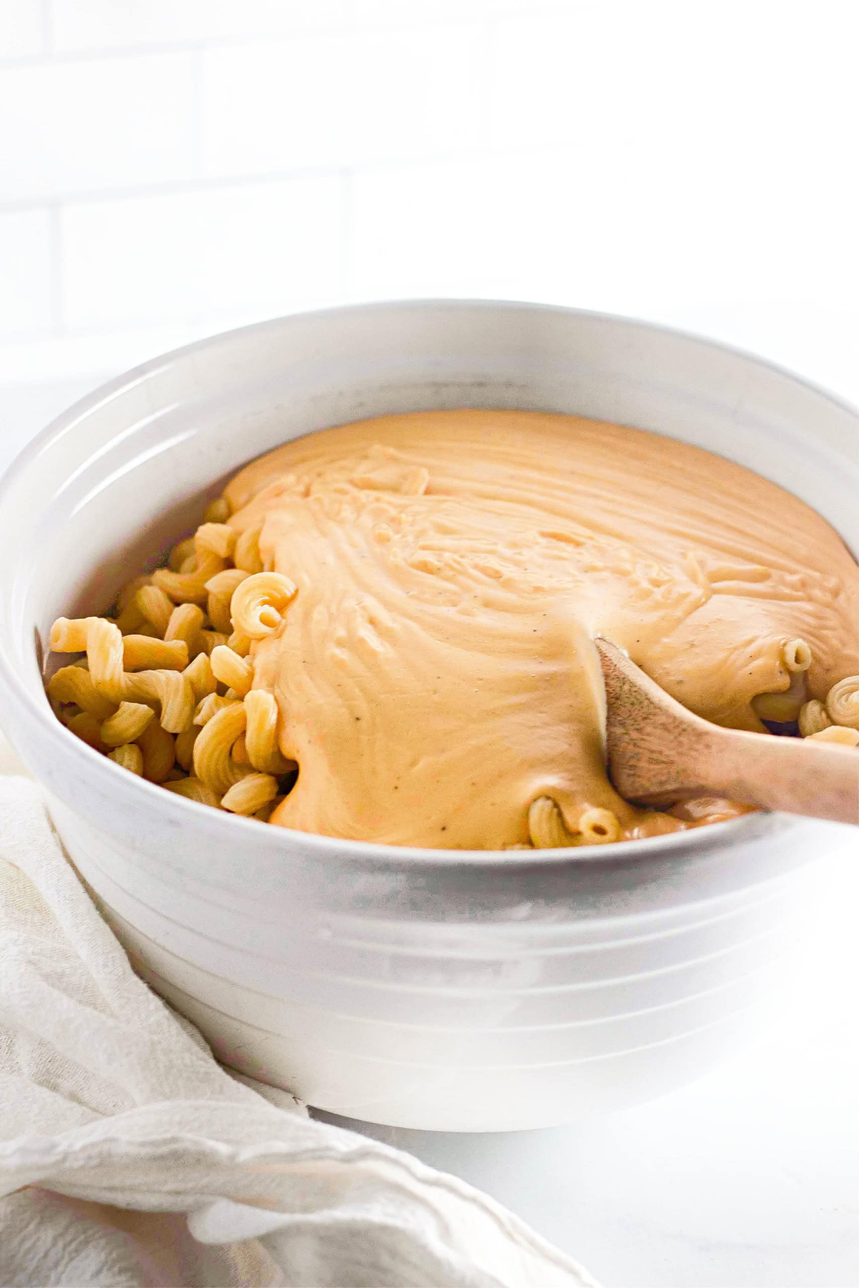 The noodles being combined with the cheese sauce in white bowl.