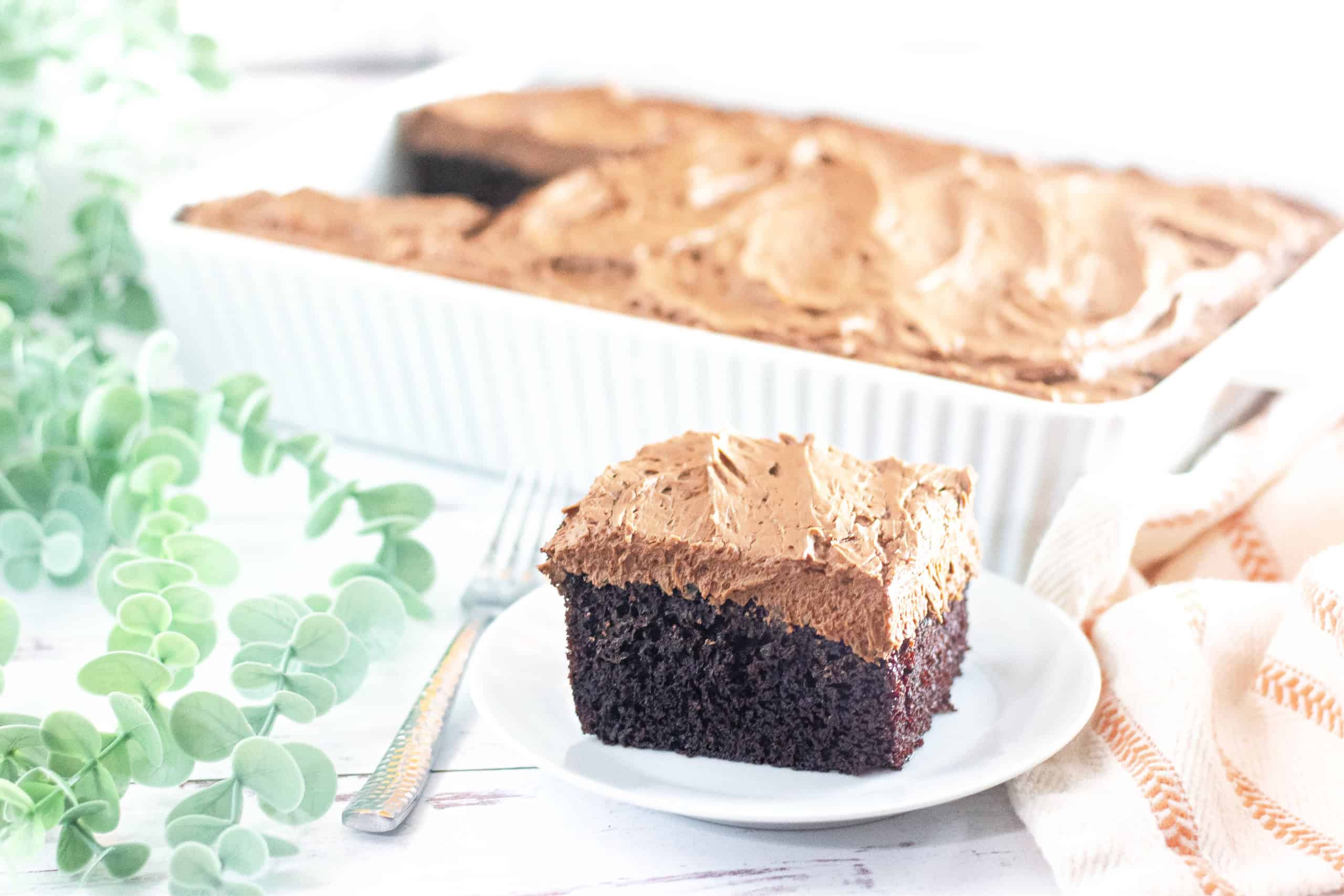 A piece of gluten free chocolate cake on white plate in front of baking pan with chocolate cake.