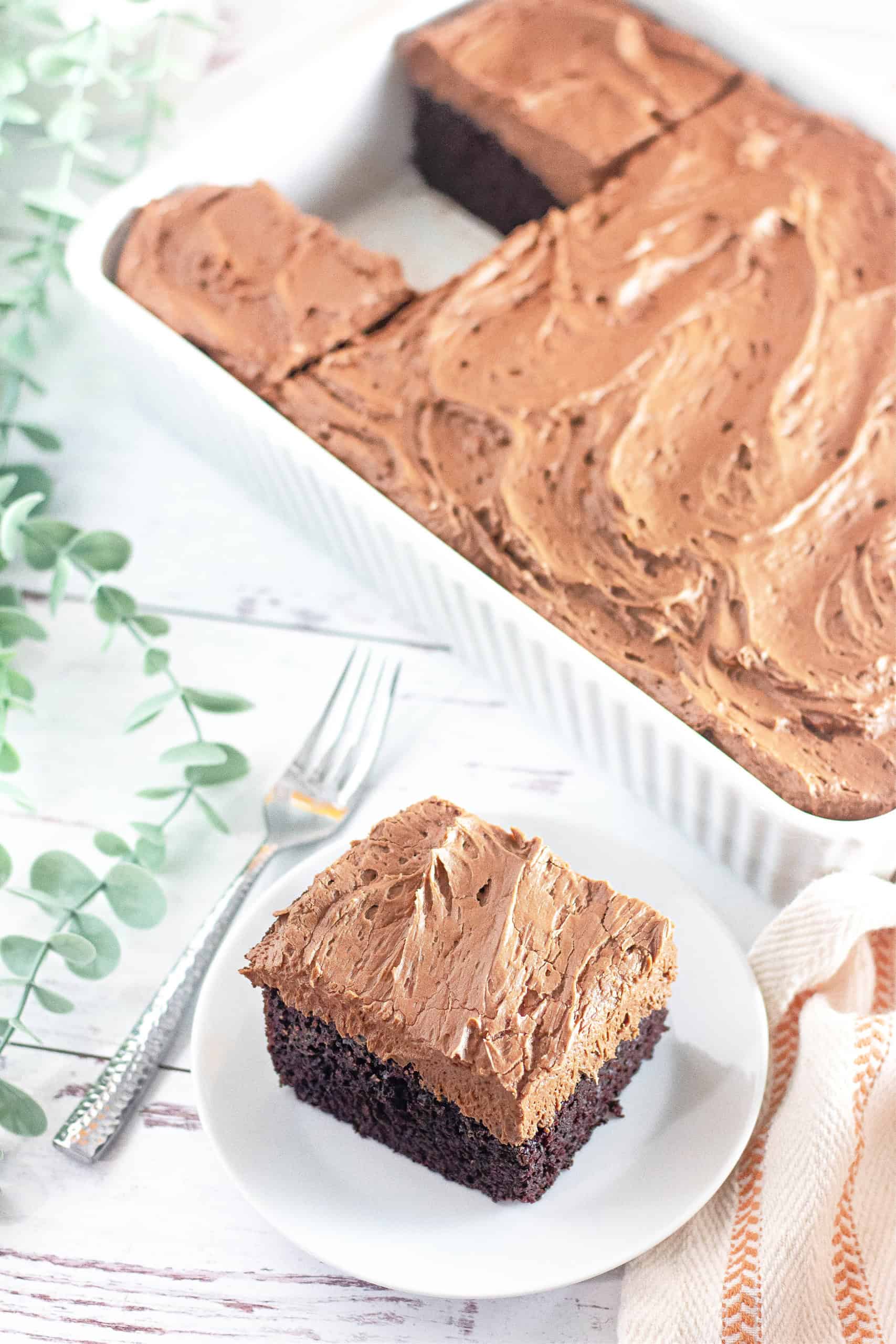 A piece of gluten free chocolate cake with chocolate buttercream frosting next to the pan of chocolate cake showing the piece missing.