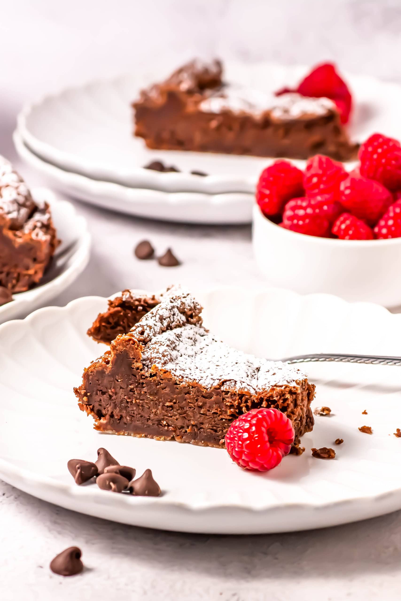 A slice of flourless chocolate torte on a white plate with another plate and slice behind it with a cup of raspberries.