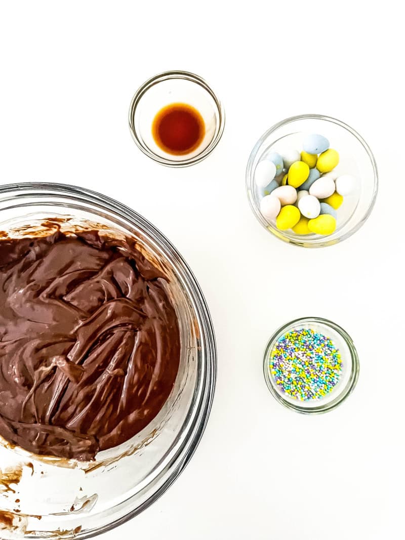Melted chocolate with the condensed milk in bowl with vanilla extract, mini egg chocolates and sprinkles.