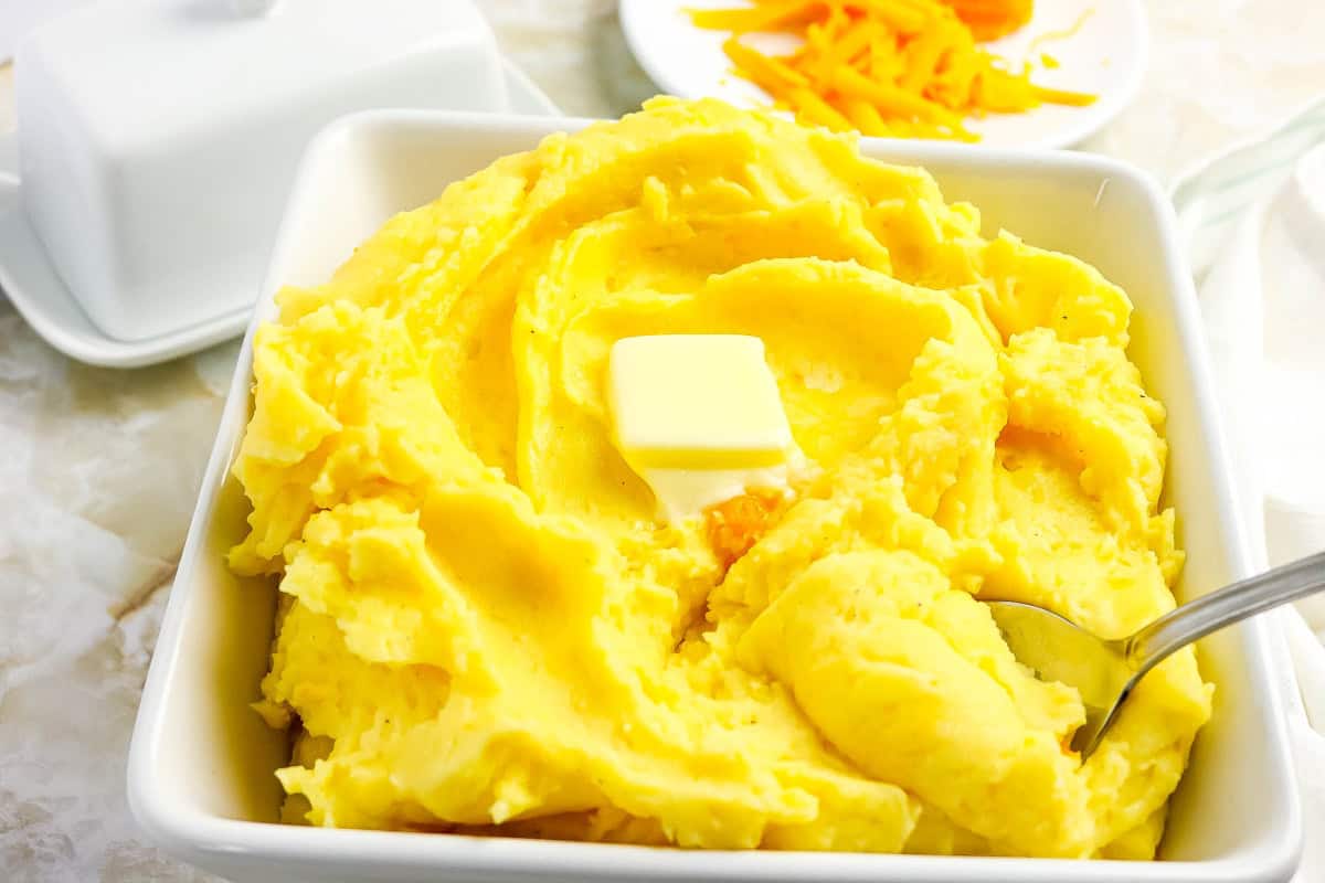 Bowl with cheesy mashed potatoes with butter on top and plate of shredded cheese beside.
