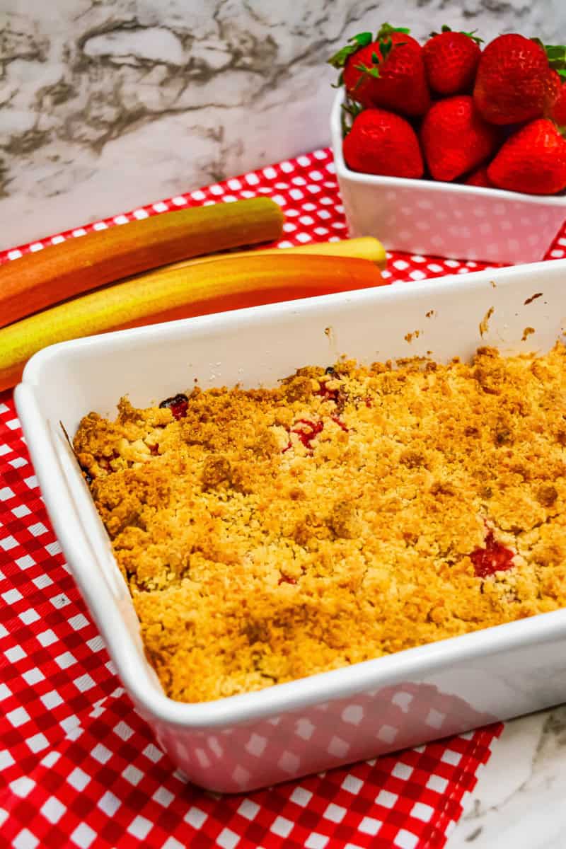 Gluten free strawberry rhubarb crumble baked on red and white checkered towel with rhubarb and strawberries next too.