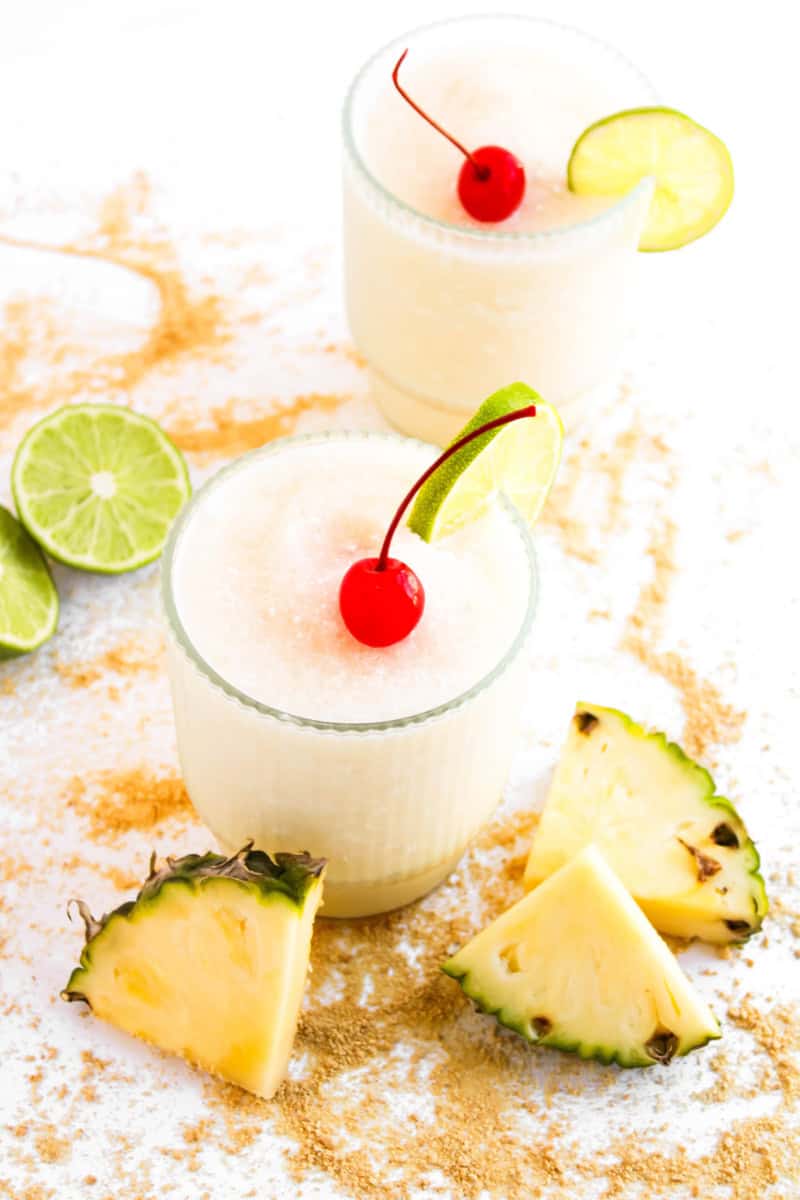 Two pina colada mocktail drinks with slices of lime and pineapple on the side and cherry on top