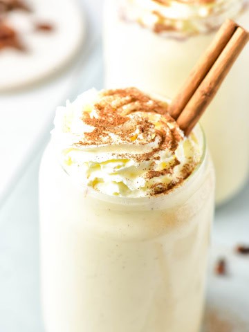 Close up of top of glass of eggnog protein shake that is topped with whipped cream and a cinnamon stick.