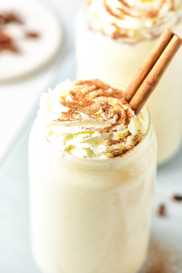 Close up of top of glass of eggnog protein shake that is topped with whipped cream and a cinnamon stick.