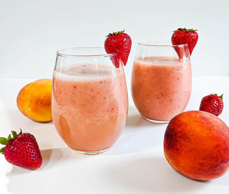 Two strawberry peach smoothies in glasses with peaches and strawberries surrounding. A strawberry hanging off of each glass.