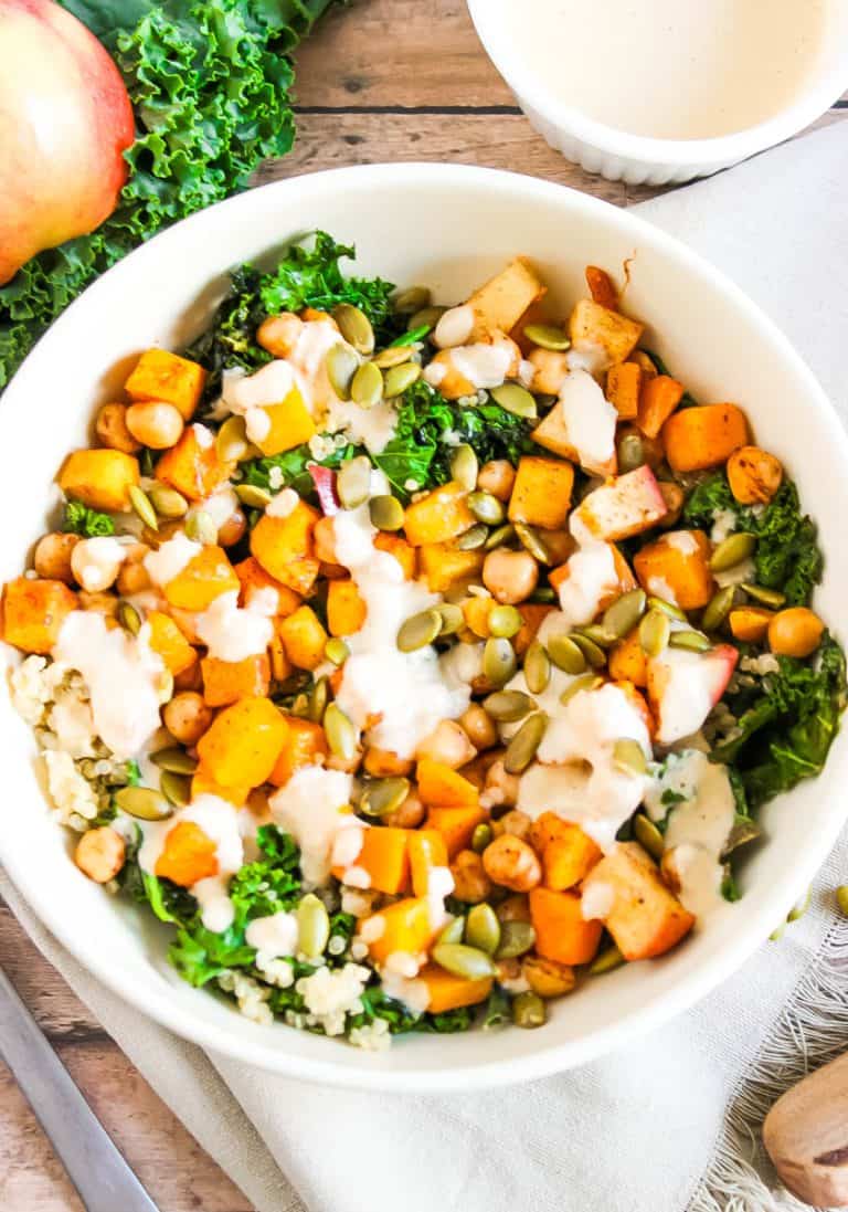 White bowl with kale, butternut squash, apples, and pumpkin seeds with dressing overtop.