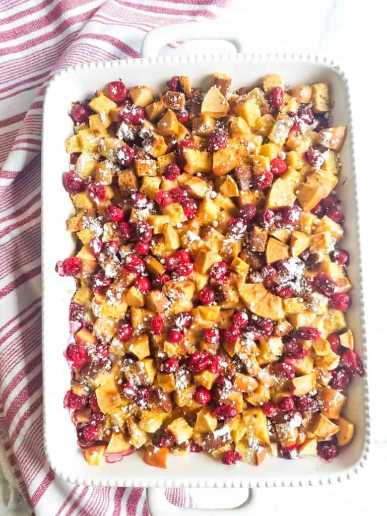 White casserole dish with gluten free cranberry apple french toast bake in it surrounded by a white and red stripped towel