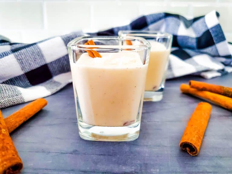 Two glasses with coquito on dark gray board with black and white towel in background surrounded by cinnamon sticks