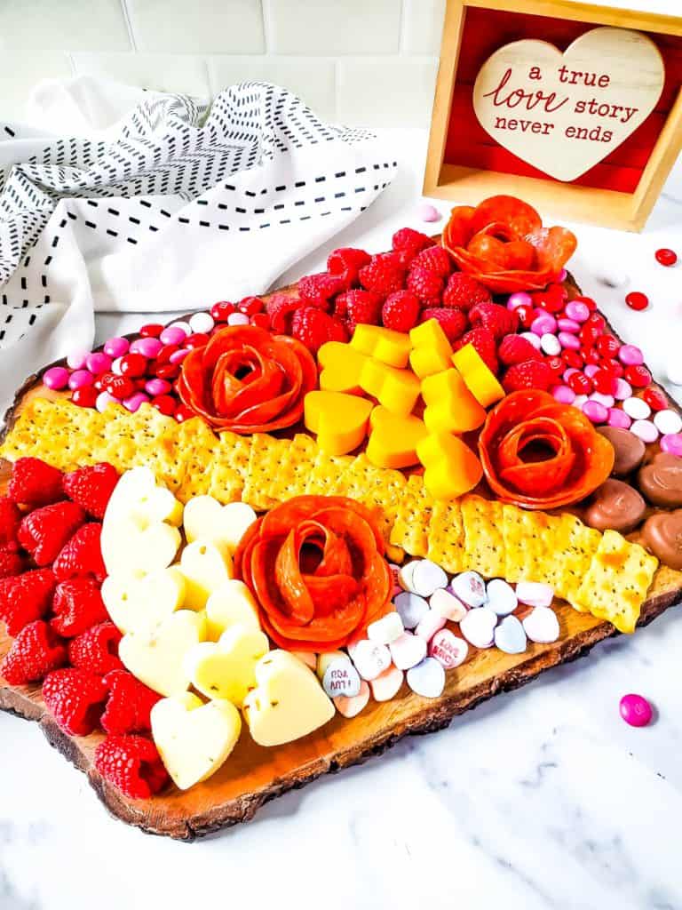 Valentine's Day Charcuterie board on white backdrop with raspberries, cheeses, crackers, candies and rose shaped pepperoni