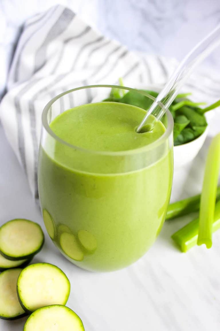 Green protein smoothie in clean glass with clear straw on white backdrop. Cucumber slices, celery sticks and spinach surround glass.