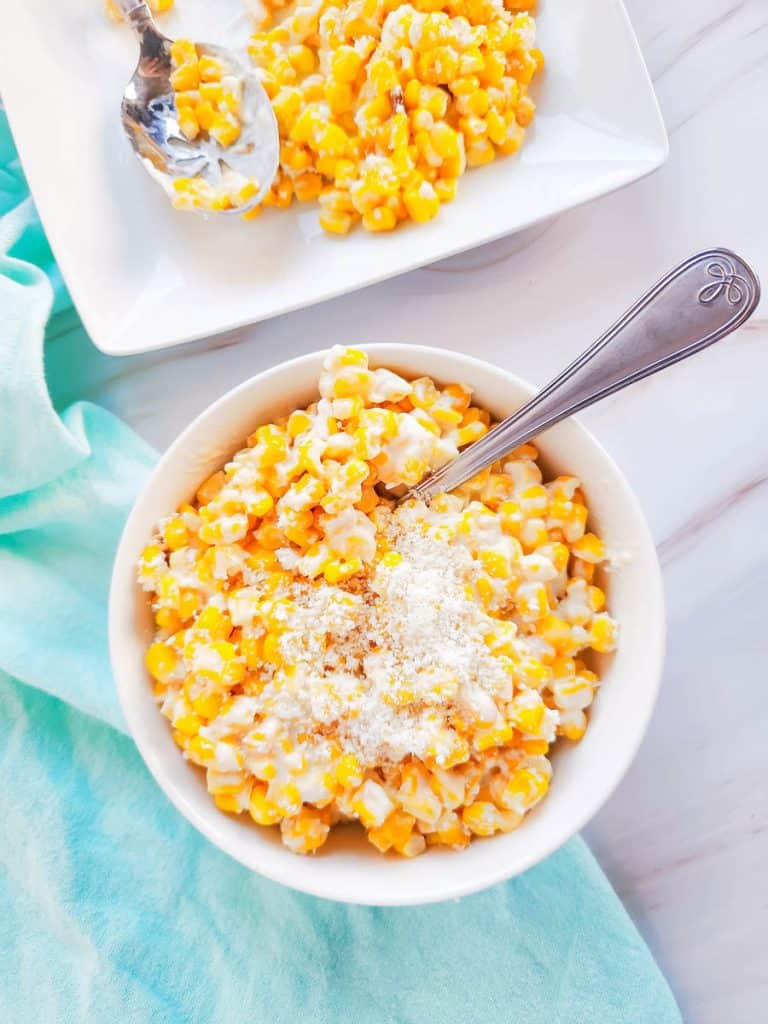 Mexican Street corn in a white bowl with a spoon. A teal towel is placed beside along with the edge of a white plate with more corn next to it.