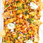 Sheet pan of bbq chicken nachos with sour cream, cheese,, bbq sauce, red onions, parsley and chips.