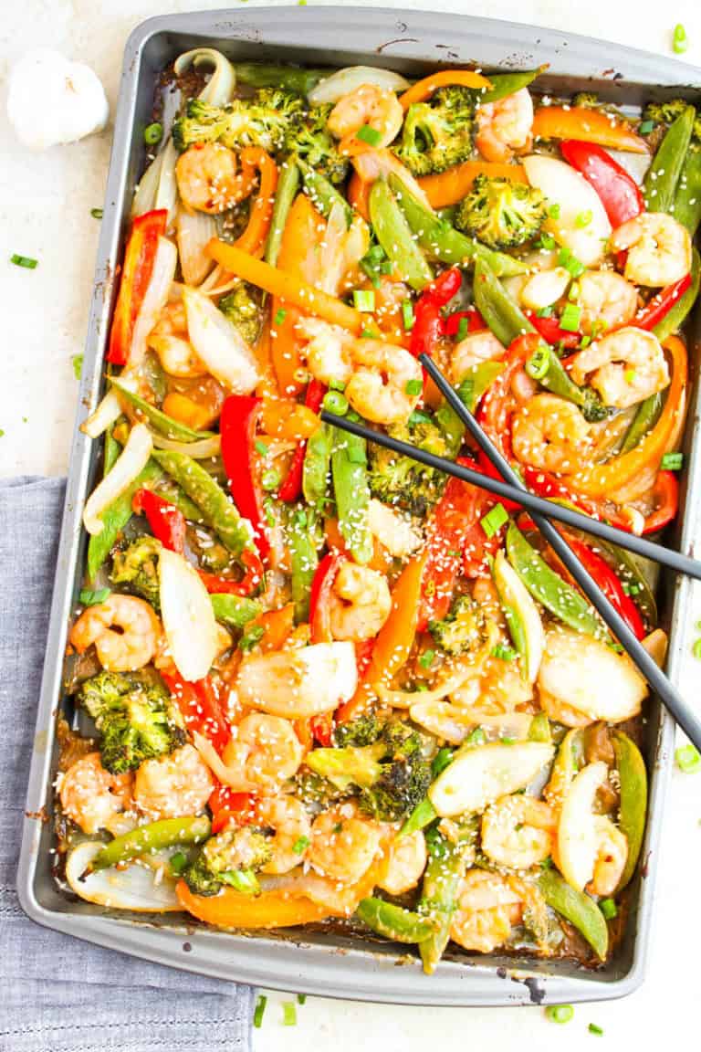 Shrimp and vegetables on one sheet pan with black chopsticks across it. Blue napkin underneath pan on white background.