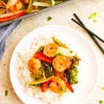 Plate with rice and sheet pan ingredients-shrimp, peppers, onion and broccoli on top. Black chopsticks next to plate with sheet pan in the top left corner on white background.