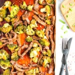 Sheet pan with beef and broccoli along with marinade sauce on white background. A silver fork and knife are next to pan. A few pieces of chopped green onion and bowl of rice in right corner of picture.