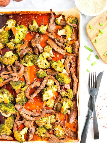 Sheet pan with beef and broccoli along with marinade sauce on white background. A silver fork and knife are next to pan. A few pieces of chopped green onion and bowl of rice in right corner of picture.